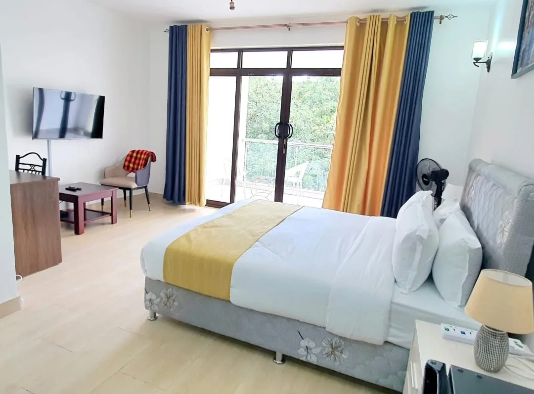 One-Bedroom Furnished – queen bed, TV, seating, and floor-to-ceiling doors to balcony